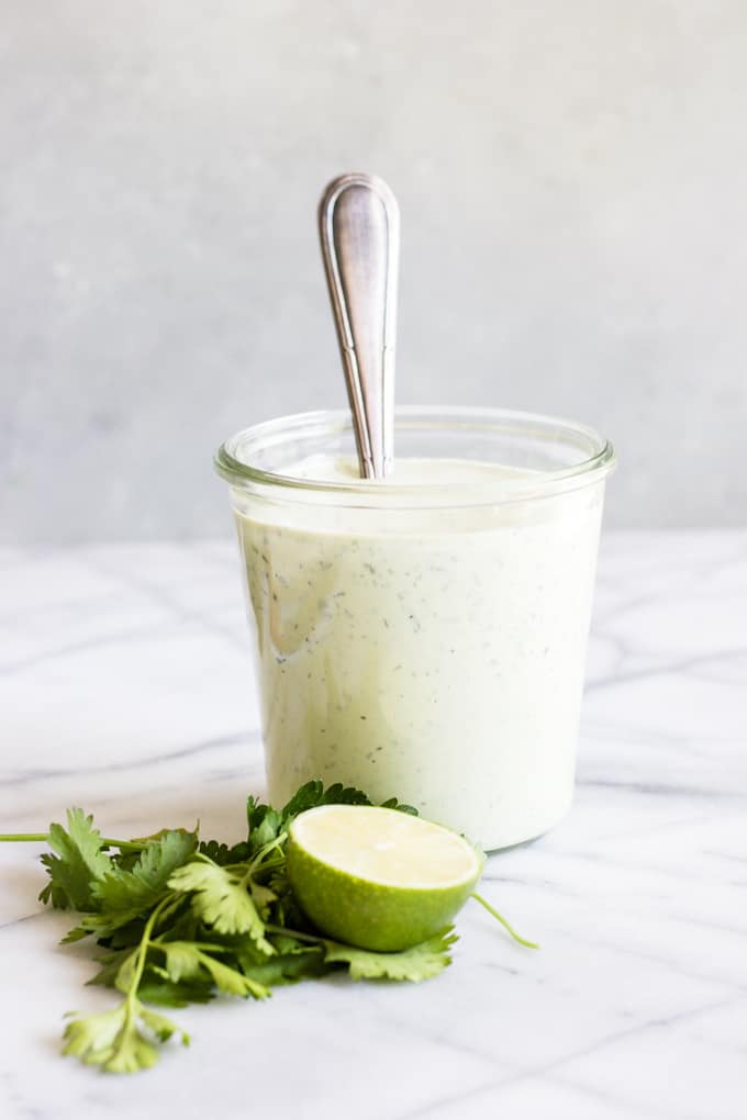 Un pot rempli de vinaigrette ranch à l'avocat dans lequel on trempe une cuillère. Entourée d'herbes fraîches et d'un demi-citron vert.