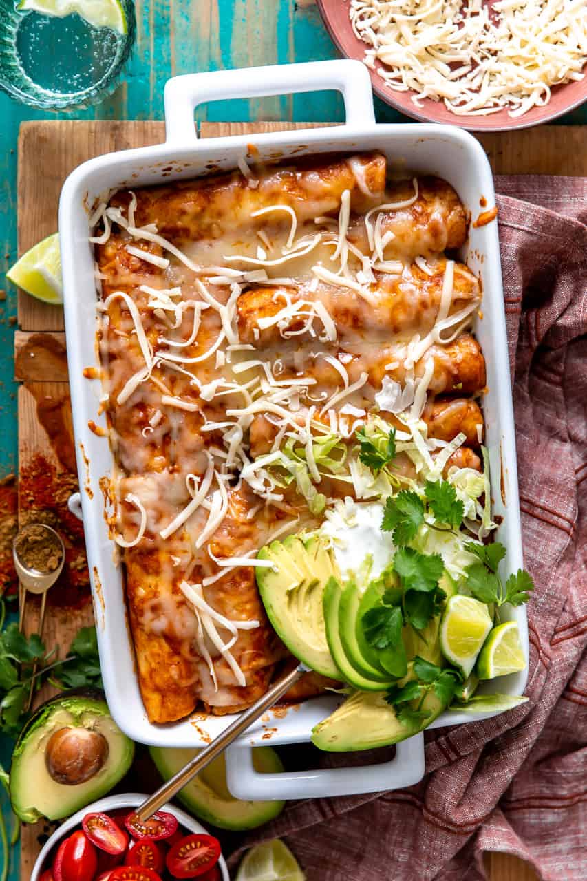 Plat à four blanc rempli d'enchiladas au poulet et garni de tranches d'avocat, de crème aigre et de coriandre fraîche.