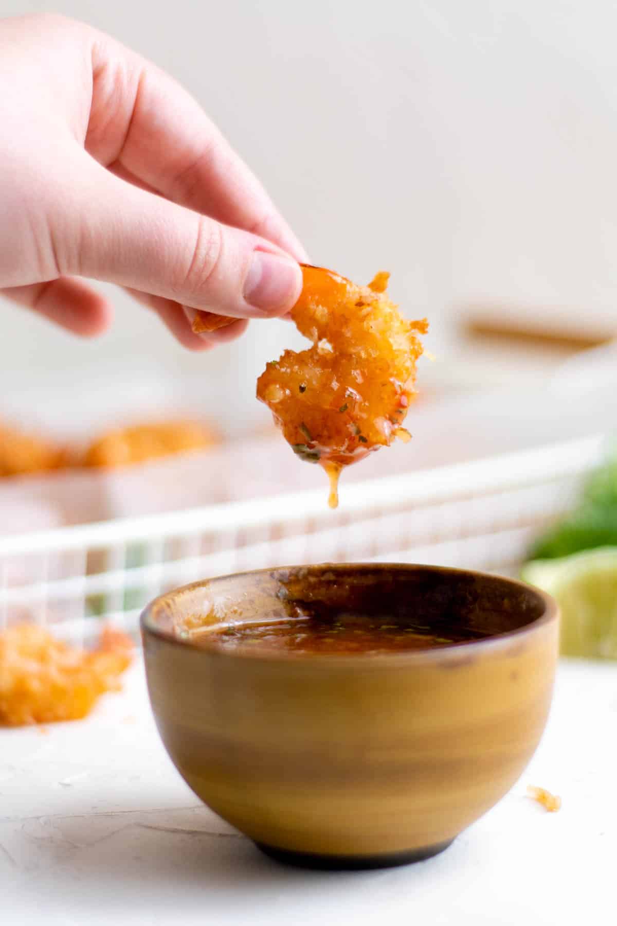 une main plongeant des crevettes croustillantes à la noix de coco dans un petit bol de sauce chili sucrée