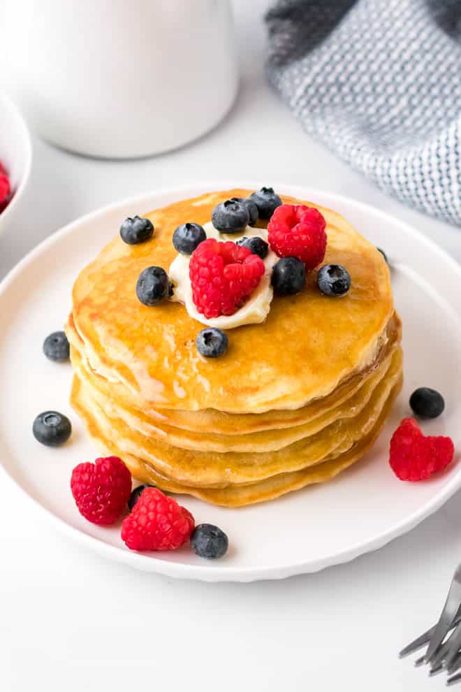 une pile de quatre crêpes maison moelleuses sur une assiette blanche avec du beurre, du sirop, des myrtilles et des framboises