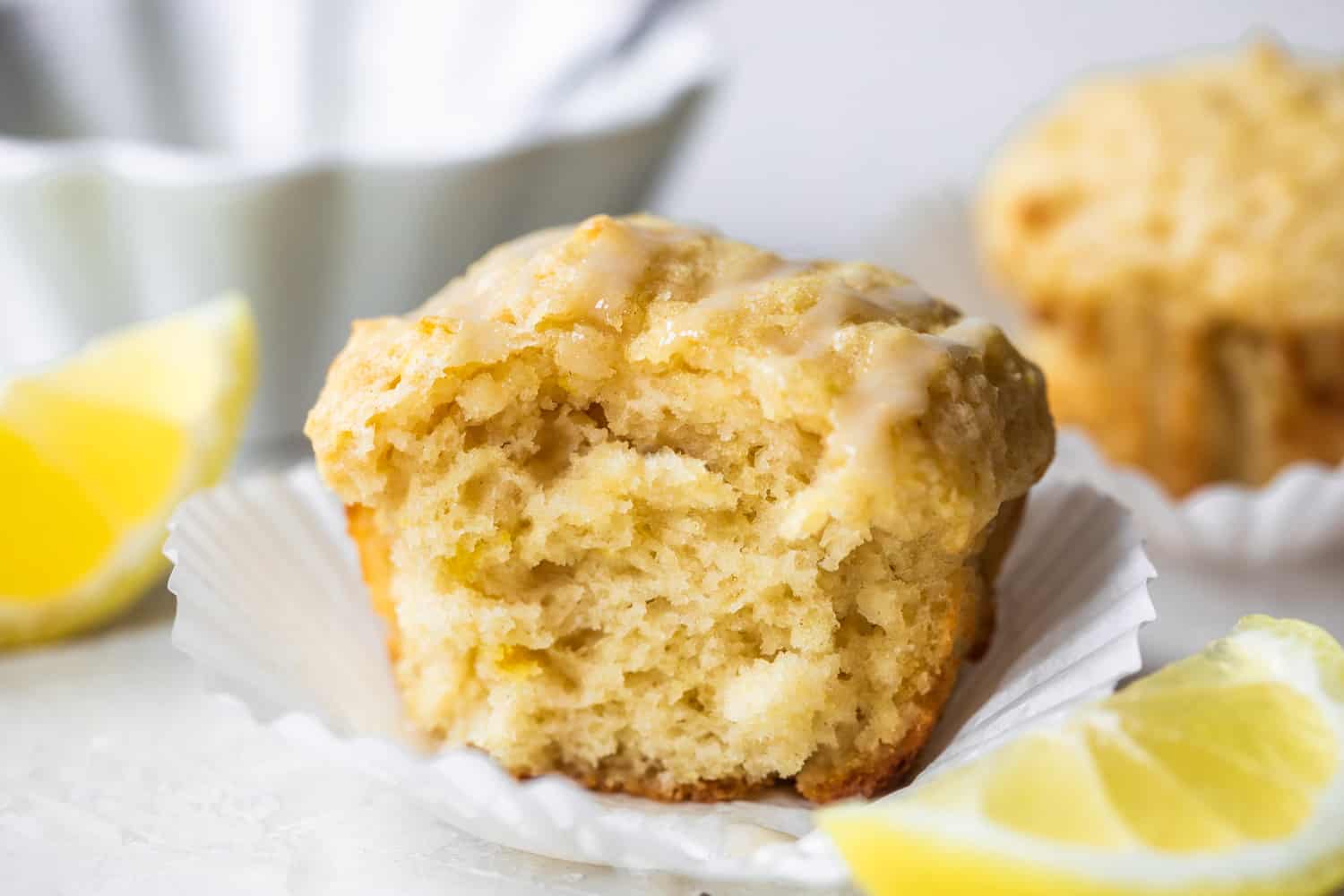 Image horizontale d'un muffin au citron cuit avec une bouchée manquante montrant une texture moelleuse. Des quartiers de citron sur le côté et un bol de glaçage derrière le muffin.