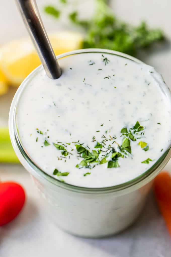 Bocal rempli de vinaigrette ranch crémeuse maison, agrémentée d'herbes hachées. Une cuillère est trempée dans la sauce ranch et des légumes frais sont disposés sur le comptoir autour du pot.