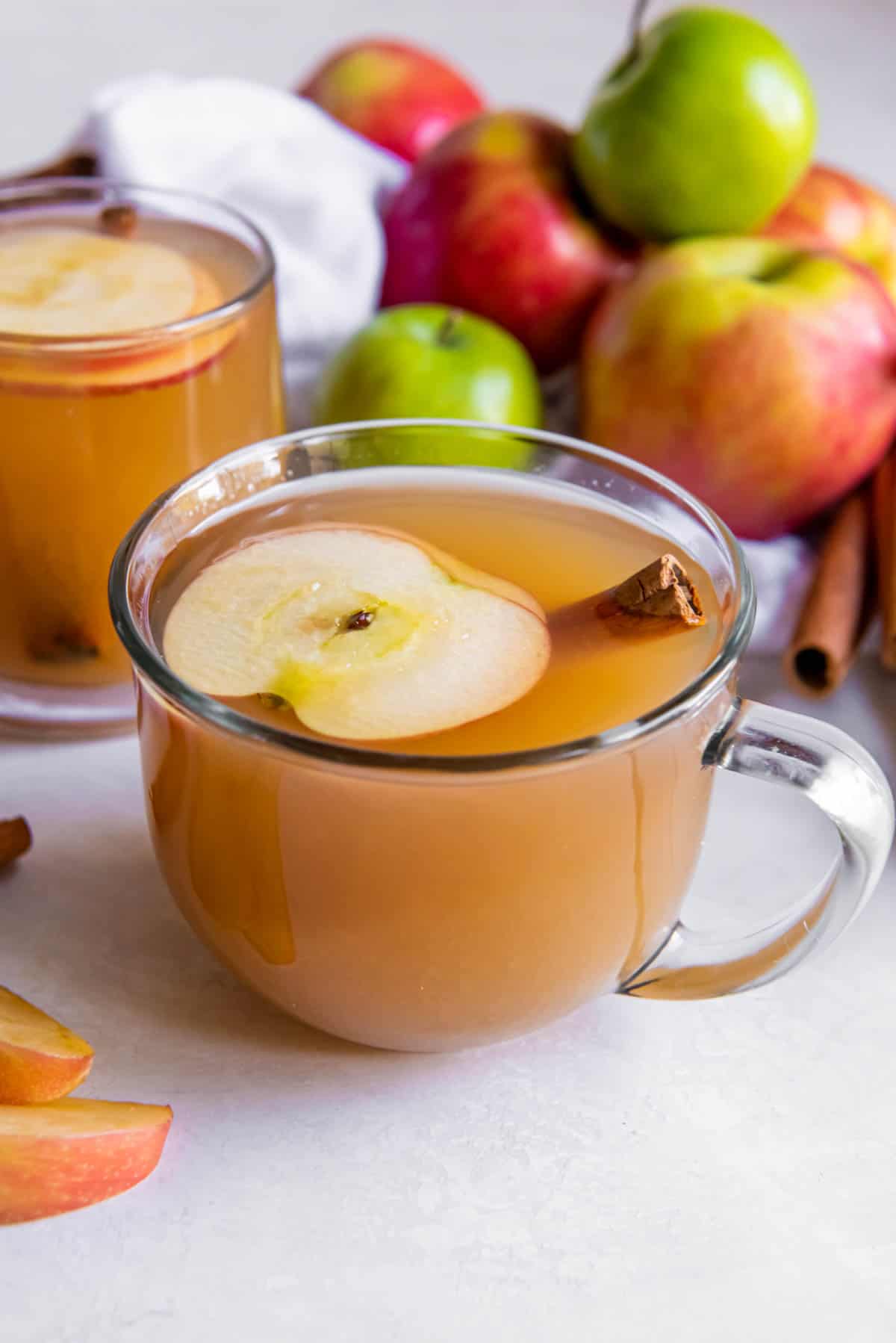 Deux verres de cidre de pomme posés sur un comptoir blanc. Une tranche de pomme et un bâton de cannelle se trouvent dans le verre de cidre.
