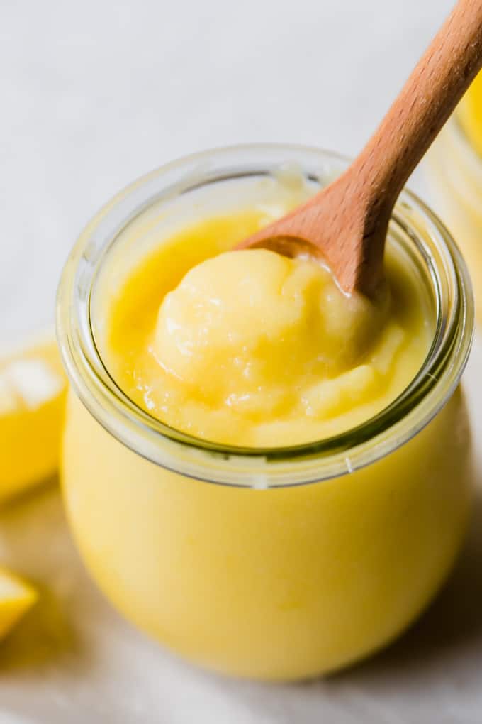 Bocal en verre tulipe rempli de lemon curd maison jaune vif avec une cuillère en bois pour le prélever.