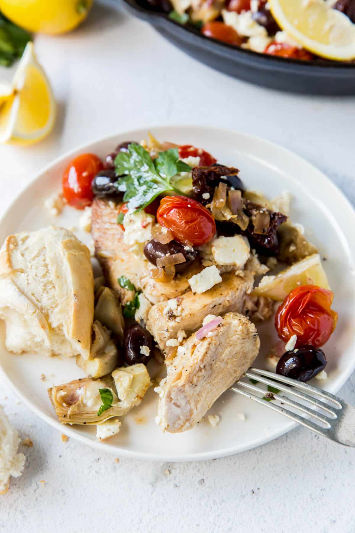 un morceau de poulet sur une assiette avec des tomates, des olives, de la feta et des artichauts sur le dessus
