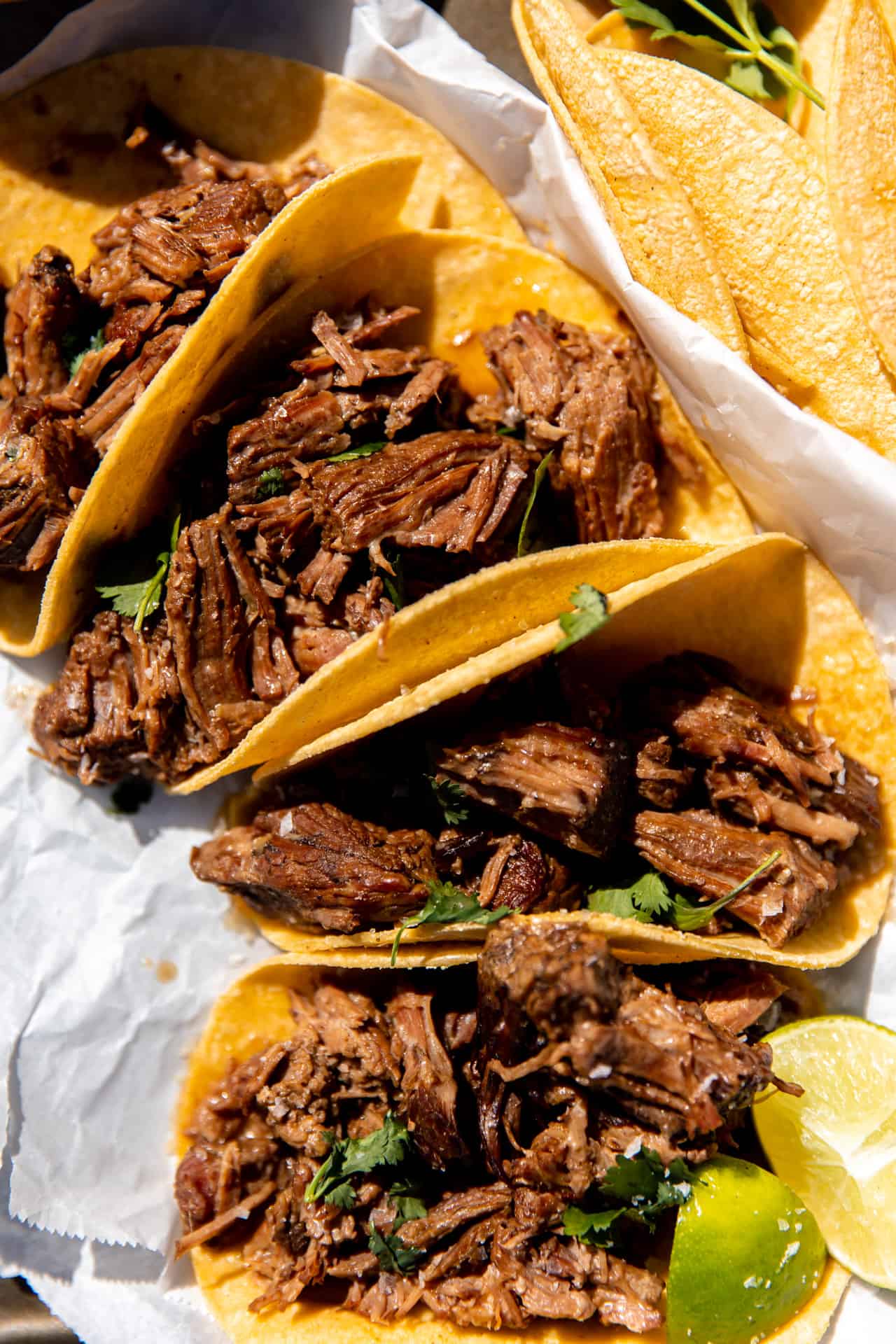 Tacos de bœuf effiloché disposés sur le comptoir au soleil avec des quartiers de citron vert et de la coriandre fraîche.
