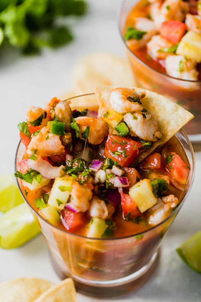 Coupe remplie de ceviche de crevettes, montrant le jus rouge, les morceaux de crevettes, le concombre, la tomate, le jalapeno, l'oignon rouge et la coriandre. Quartiers de citron vert sur le côté, chips de tortilla et coriandre.