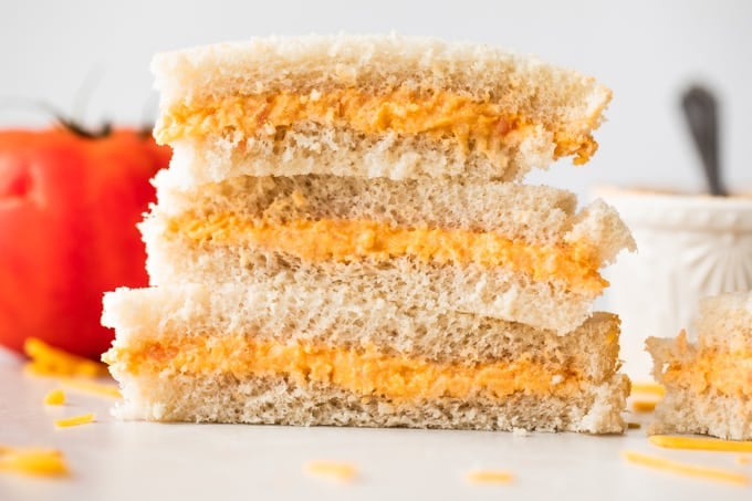 Pile de sandwichs à la tomate sur du pain blanc, avec du fromage râpé sur le comptoir autour d'eux, une tomate en arrière-plan et un ramequin rempli de tomates à la crème.