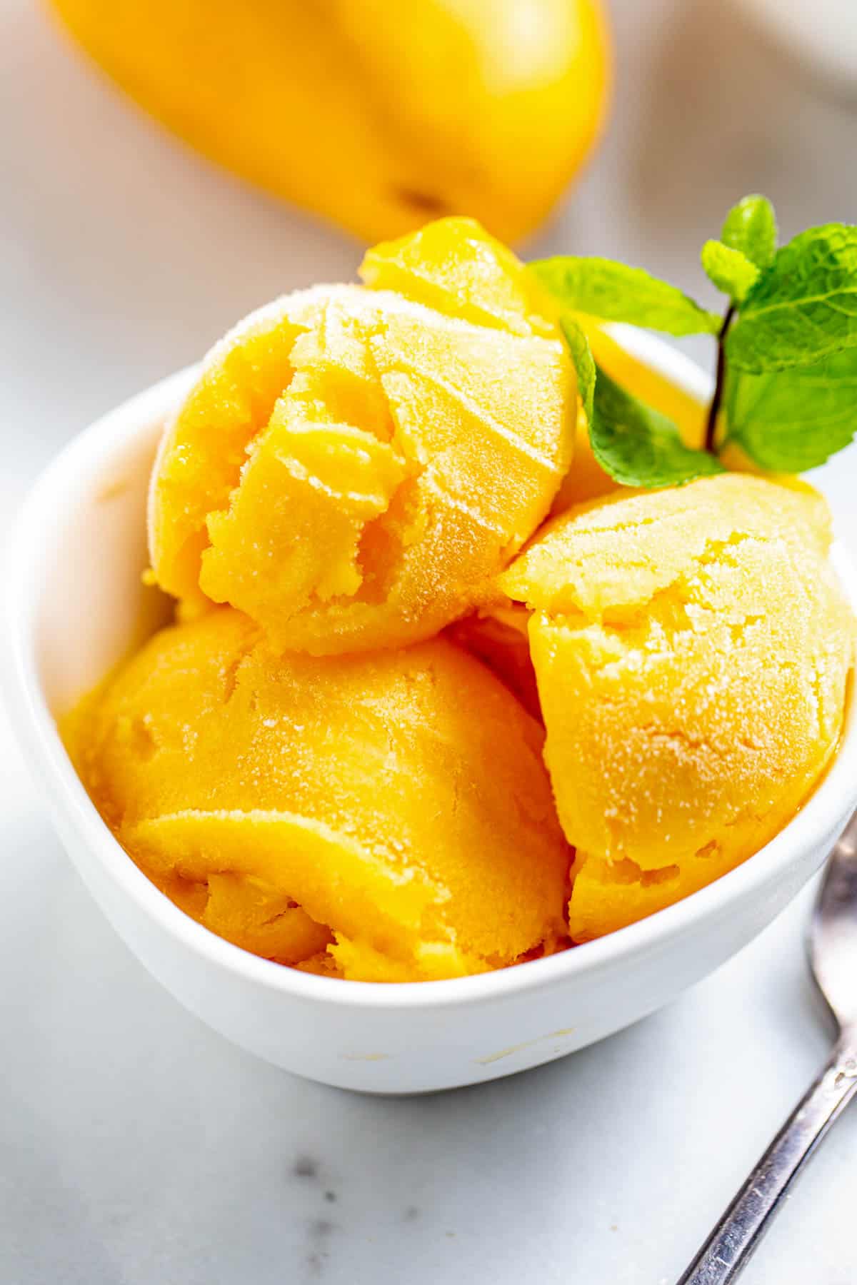 Trois boules de sorbet à la mangue fait maison dans un bol blanc avec un brin de menthe poivrée sur le dessus.