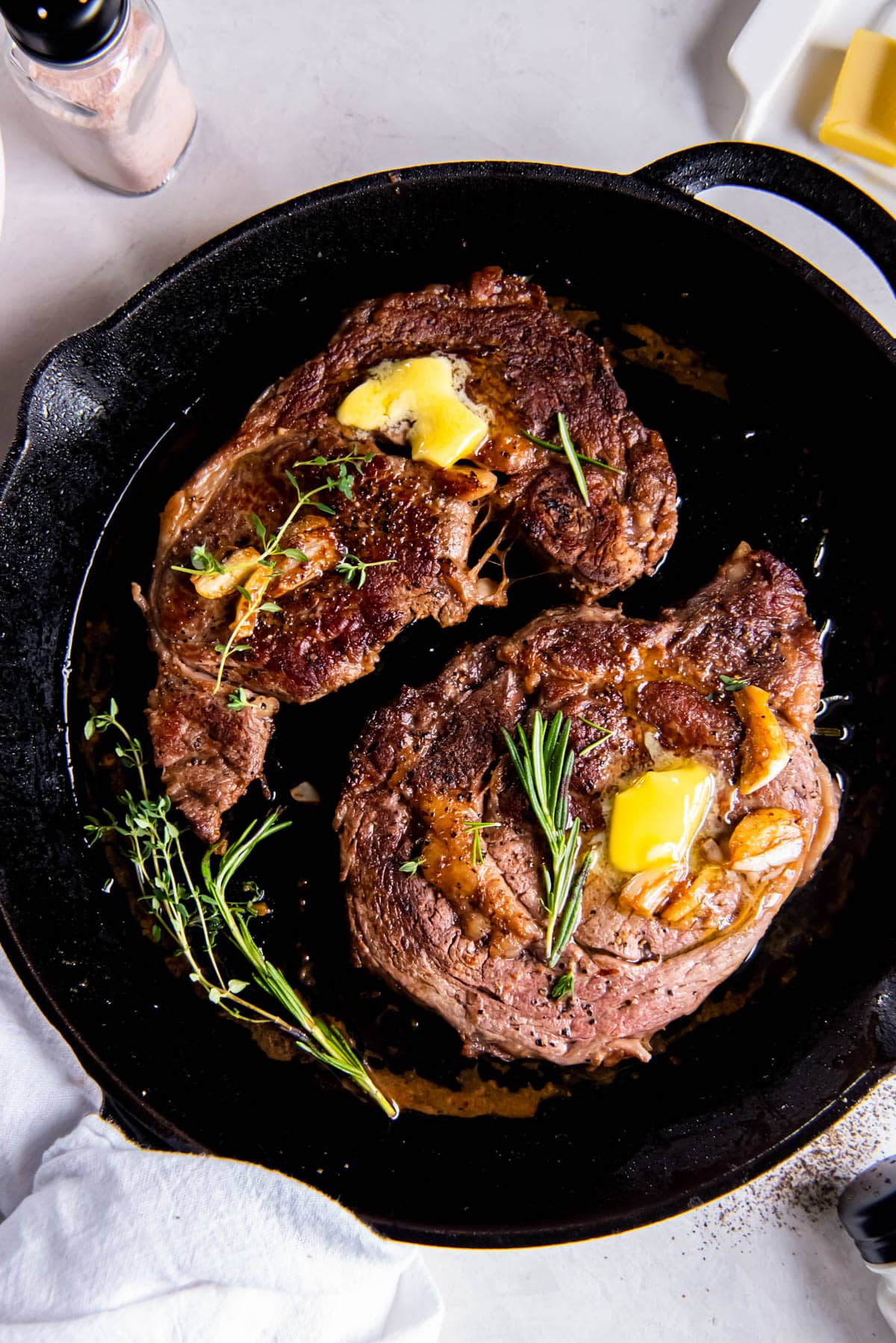 Deux steaks de côte de bœuf saisis dans une poêle en fonte avec une tranche de beurre fondu et du romarin frais sur le dessus.