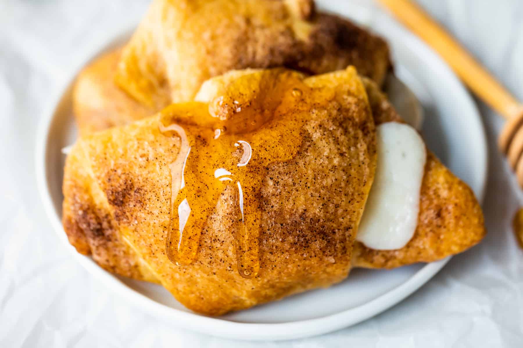 Rouleaux de cheesecake sopapilla cuits au four sur une assiette blanche avec un filet de miel qui coule, un bâtonnet de miel sur le côté.