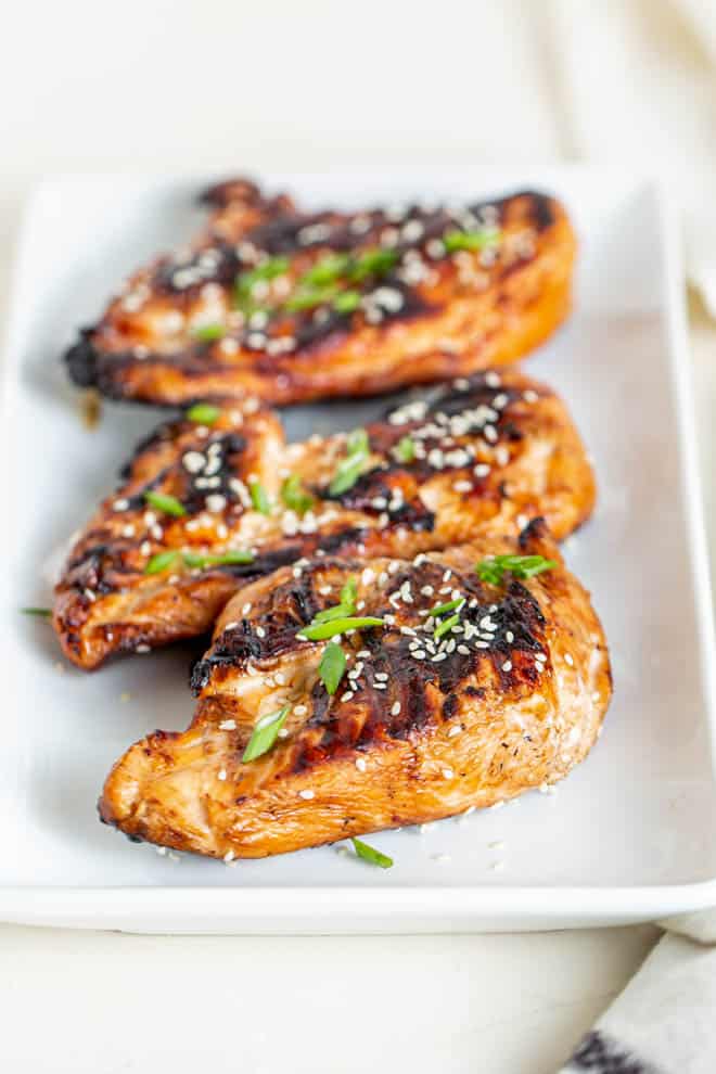 trois morceaux de poulet teriyaki grillé posés sur une assiette blanche et garnis de graines de sésame et de ciboulette