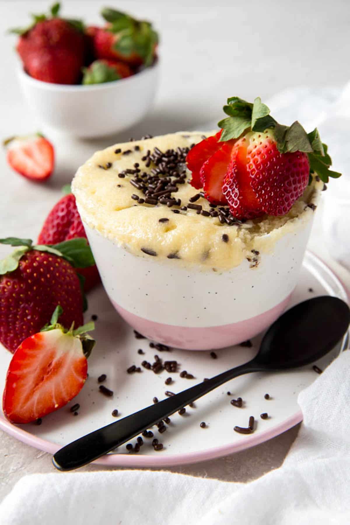 Mug cake à la vanille avec des paillettes supplémentaires et une fraise coupée en tranches sur le dessus. Une cuillère est posée sur une assiette à côté du mug cake.